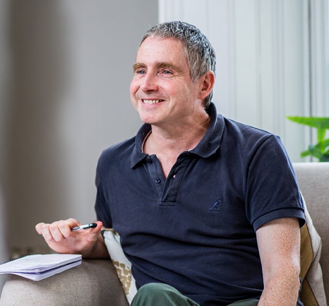 A wide photo of Martin Hawkins, smiling while sitting on a sofa