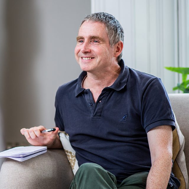 A wide photo of Martin Hawkins, smiling while sitting on a sofa