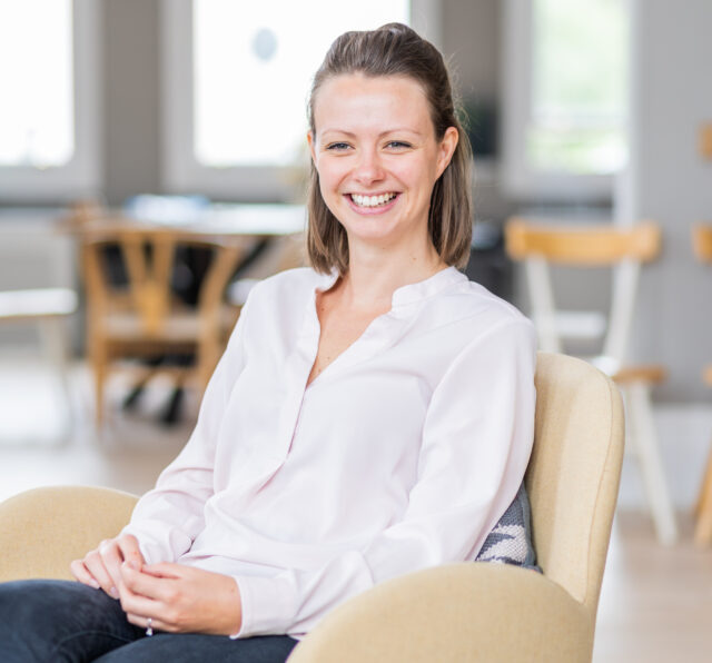 Photo of Fiona Hiller, smiling while sitting on a chair, wearing a pink shirt and jeans.