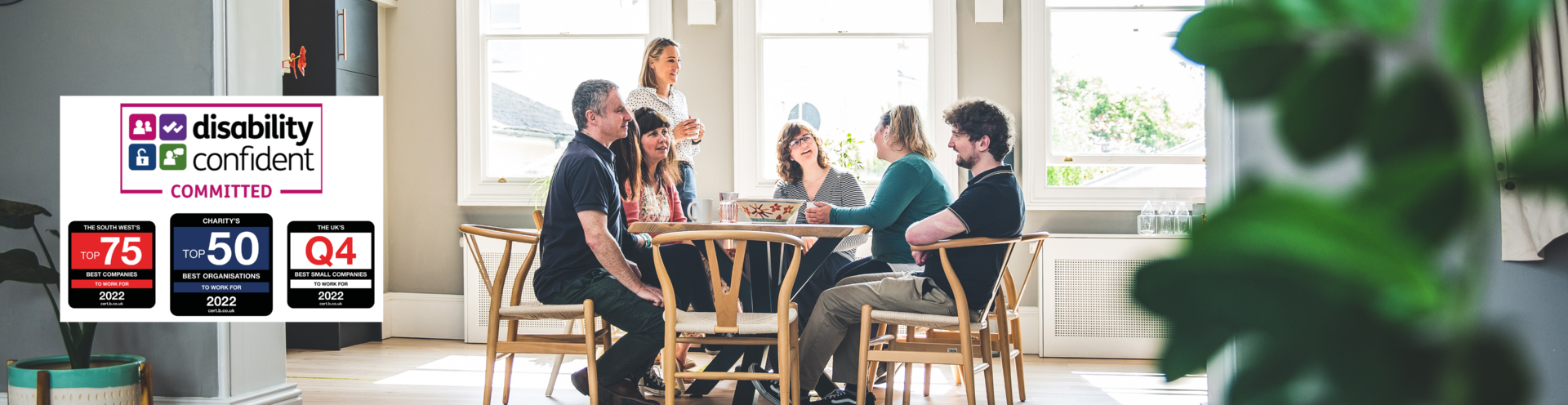Photograph of Barnwood staff, having lunch in the kitchen on a bright sunny day. There are four logos to the left side, including Disability Confident Committed, The South West Top 75 Best companies to work for, Q3 2022, Charity's Top 25 Best Organisations to work for, Q3 2022 and The UK's Top 100 Best Small Companies to work for, Q4 2022.