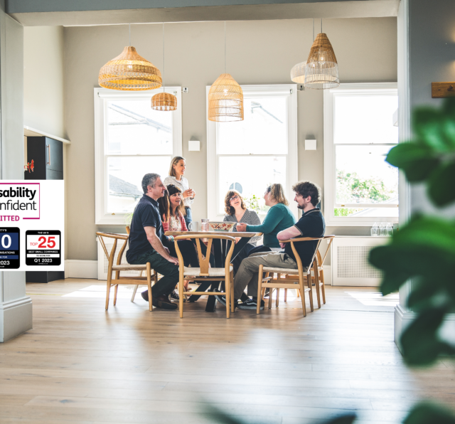 Photograph of Barnwood staff, having lunch in the kitchen on a bright sunny day. There are four logos to the left side, including Disability Confident Committed, The South West Top 25 Best companies to work for, Q1 2023, Charity's Top 10 Best Organisations to work for, Q1 2023 and The UK's Top 25 Best Small Companies to work for, Q1 2023.