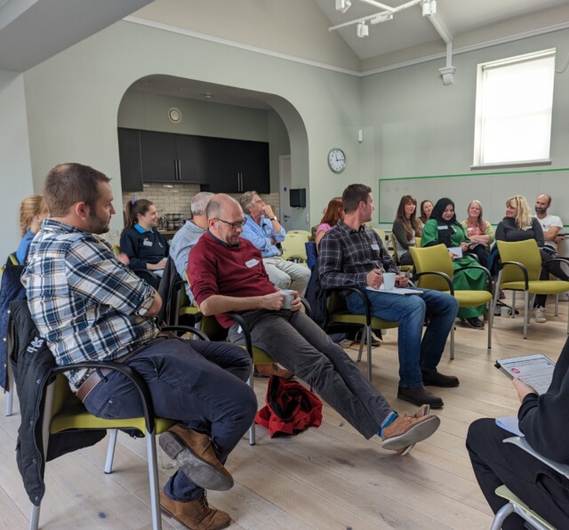 Photo of participants at a Barnwood Trust learning event. People are sat in rows of chairs listening to a presenter.