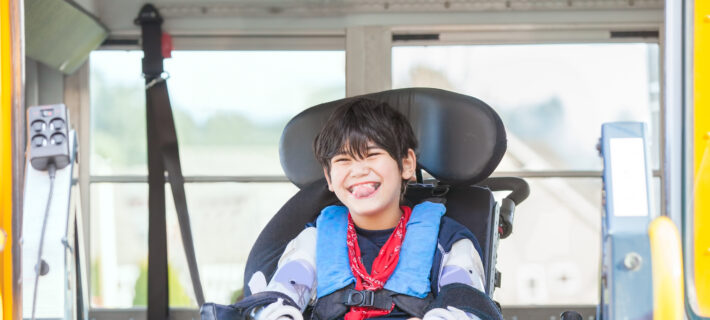 Happy biracial little boy with special needs sitting in wheelchair, riding on yellow school bus lift, going to school