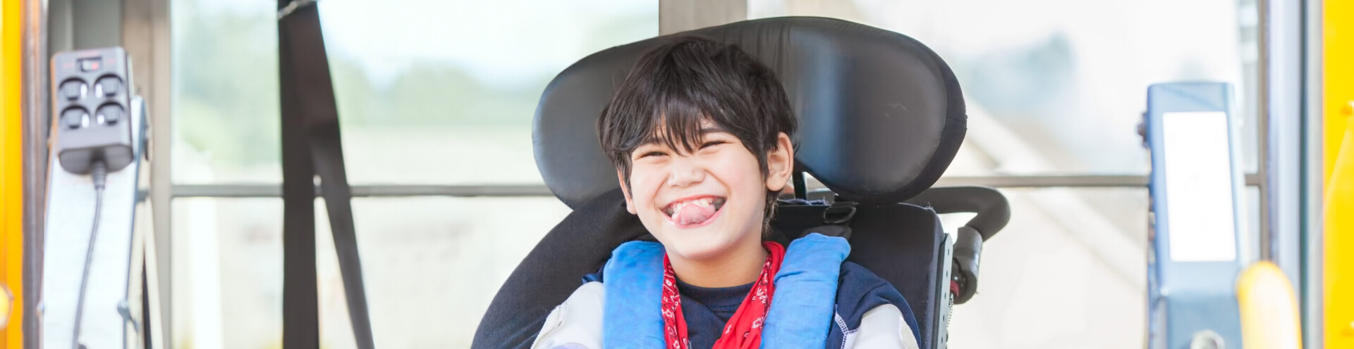 Happy biracial little boy with special needs sitting in wheelchair, riding on yellow school bus lift, going to school