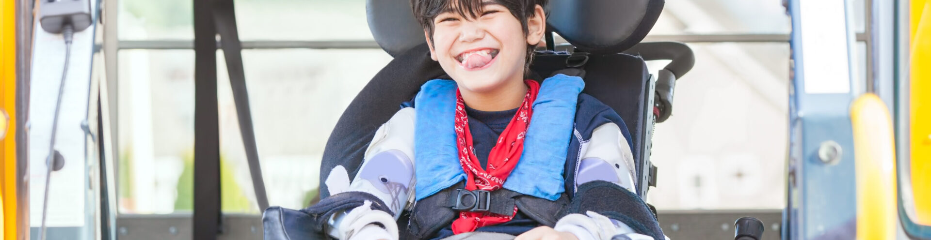 Happy biracial little boy with special needs sitting in wheelchair, riding on yellow school bus lift, going to school