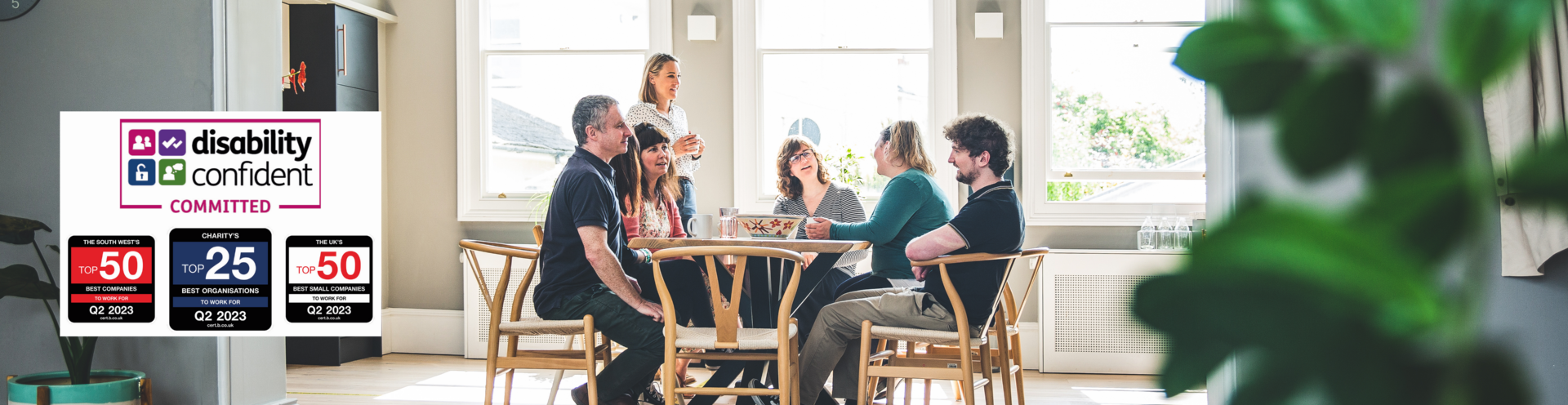 Photograph of Barnwood staff, having lunch in the kitchen on a bright sunny day. There are four logos to the left side, including Disability Confident Committed, The South West Top 25 Best companies to work for, Q2 2023, Charity's Top 10 Best Organisations to work for, Q2 2023 and The UK's Top 25 Best Small Companies to work for, Q2 2023.