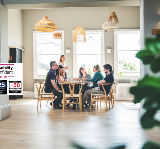 Photograph of Barnwood staff, having lunch in the kitchen on a bright sunny day. There are four logos to the left side, including Disability Confident Committed, The South West Top 25 Best companies to work for, Q2 2023, Charity's Top 10 Best Organisations to work for, Q2 2023 and The UK's Top 25 Best Small Companies to work for, Q2 2023.