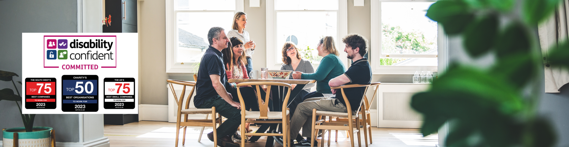 Photograph of Barnwood staff, having lunch in the kitchen on a bright sunny day. There are four logos to the left side, including Disability Confident Committed, The South West Top 25 Best companies to work for, 2023, Charity's Top 10 Best Organisations to work for, 2023 and The UK's Top 25 Best Small Companies to work for, 2023.