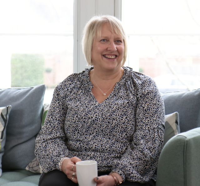 Photo of Justine Norman, sitting on a sofa with a tea cup and a smile on her face.