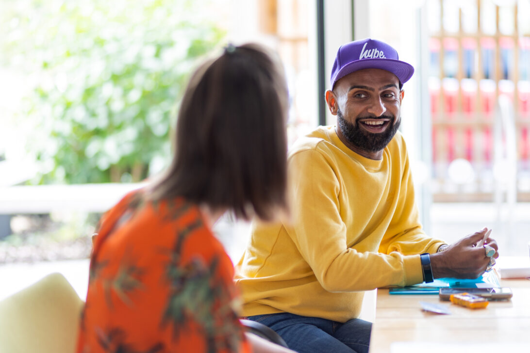 Photo of a Barnwood Circle member, smiling while sitting at a table and listening listening to a Barnwood member of staff on a bright day. 