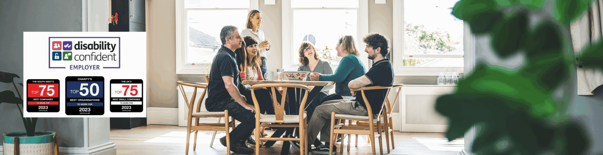 Photograph of Barnwood staff, having lunch in the kitchen on a bright sunny day. There are four logos to the left side, including Disability Confident Employer, The South West Top 25 Best companies to work for, 2023, Charity's Top 10 Best Organisations to work for, 2023 and The UK's Top 25 Best Small Companies to work for, 2023.