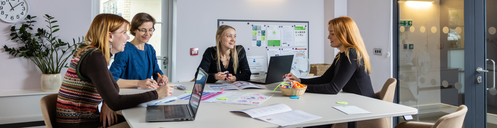 4 women talking to each other and sitting around a table with laptops and open booklets on it.
