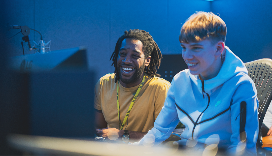 A man and a younger man sitting next to each look and smiling whilst looking at ta computer screen