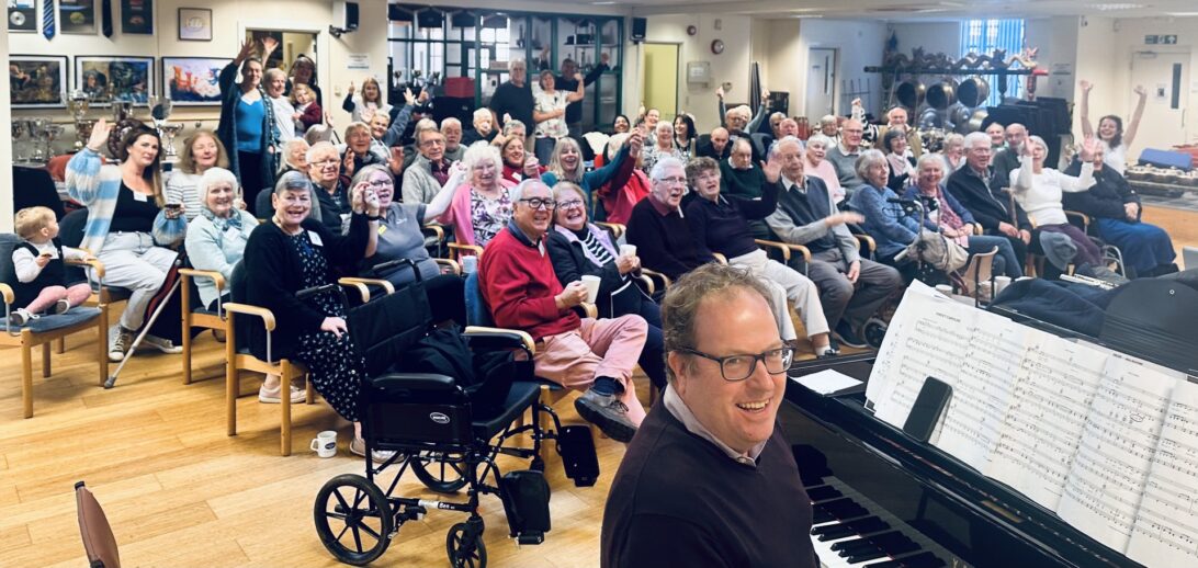 A large group of people smiling and waving behind a man sitting at a piano, all looking at the camera.