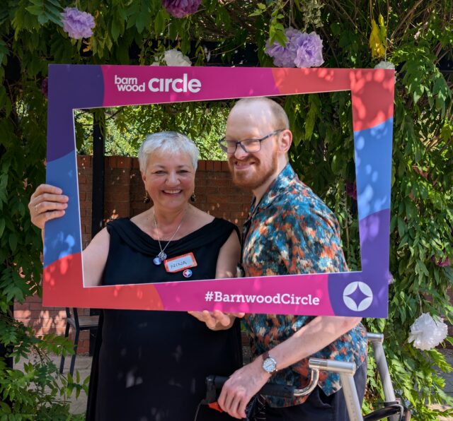 Two Barnwood Circle members smiling at the camera whilst holding a Barnwood Circle branded frame.